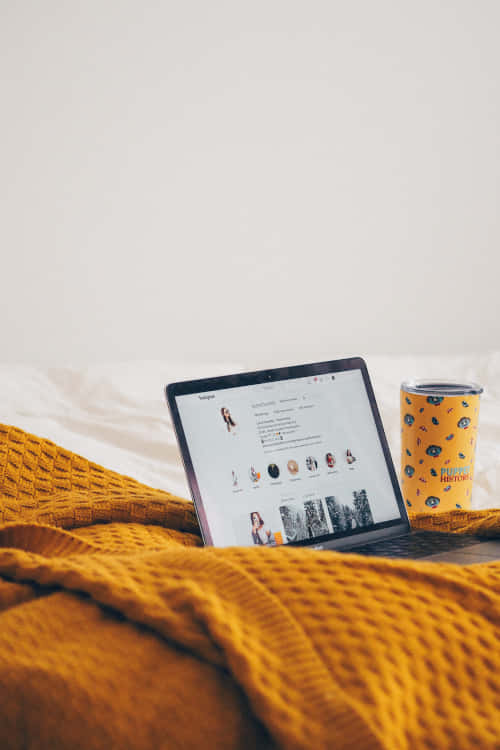 A laptop on a bed covered by a yellow bedsheet with a cup beside it